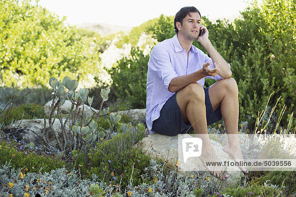 Ein erwachsener Mann  der auf einem Felsen sitzt und auf einem Handy spricht.