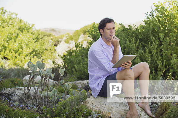 Mann sitzt auf einem Felsen mit einem digitalen Tablett und denkt nach.