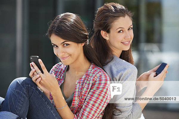 Porträt zweier junger Frauen  die sich Rücken an Rücken lehnen und Mobiltelefone benutzen