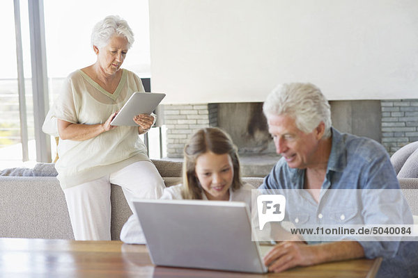 Man using a laptop with his granddaughter and his wife working on a digital tablet in background