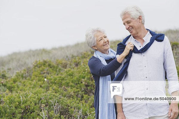 Ältere Frau  die am Strand einen Pullover auf die Schulter ihres Mannes legt.