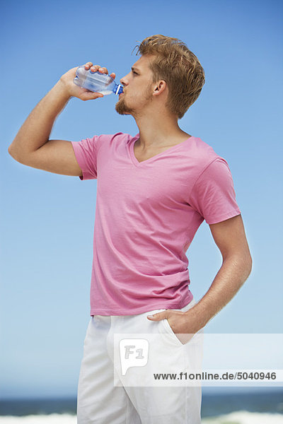Junger Mann trinkt Wasser aus einer Wasserflasche am Strand