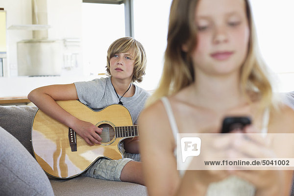 Teenager-Junge spielt Gitarre  während seine Schwester ein Handy benutzt.