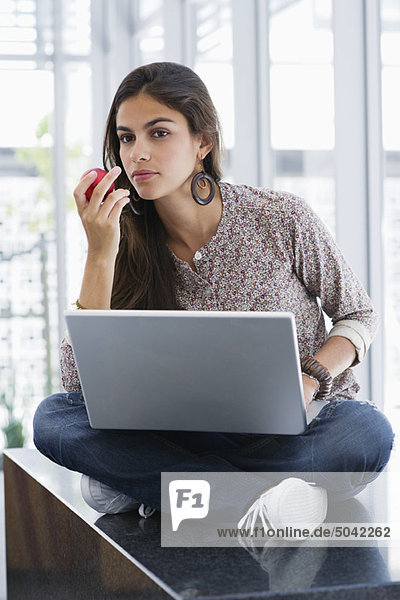 Schöne Frau  die einen Apfel isst  während sie einen Laptop benutzt.