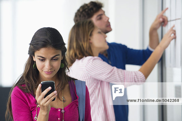 Universitätsstudenten  die im Bulletin Board nach Testergebnissen suchen  während Frauen Textnachrichten auf dem Handy abrufen.
