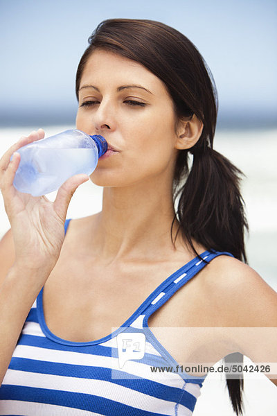 Nahaufnahme einer Frau  die Wasser aus einer Wasserflasche am Strand trinkt.
