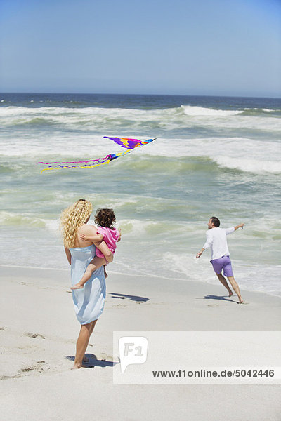 Mother carrying child while man flying kite on the beach