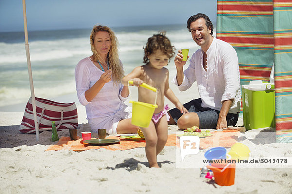 Mädchen beim Spielen am Strand mit ihren Eltern  die hinter ihr sitzen