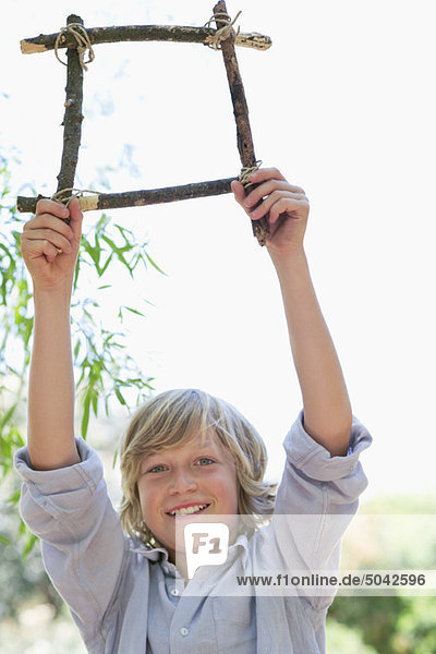 Portrait of a cute little boy holding frame of driftwood with arms raised outdoors