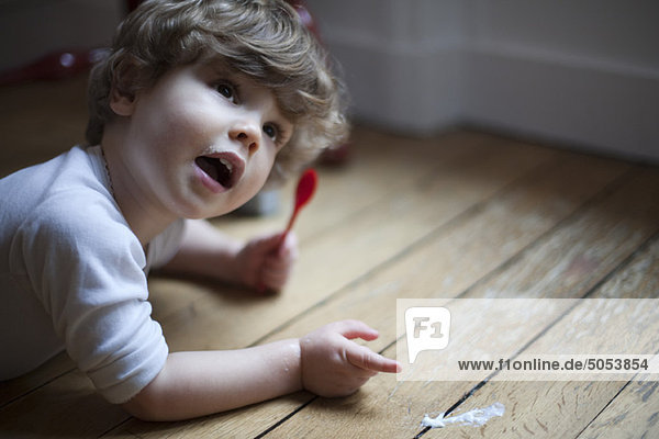 Toddler lying on floor with spoon in hand  pointing to spilled food