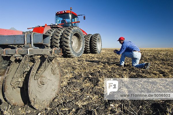 a man examines newly cultivated soil and wheat stubble beside a tractor pulling cultivating equipment near Lorette  Manitoba  Canada