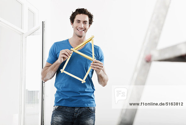 Junger Mann hält Hausmodell-Taschenlineal in renovierender Wohnung