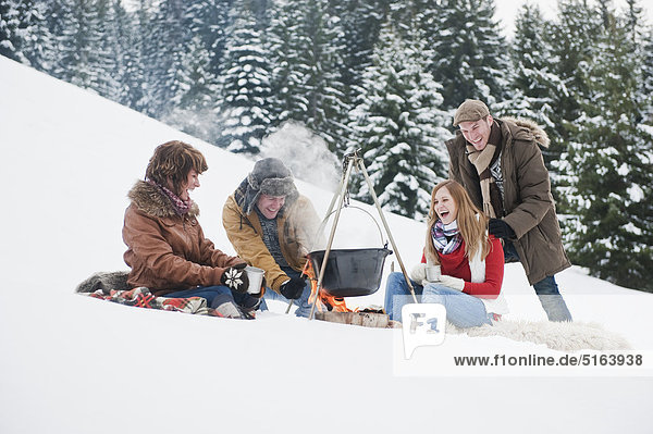 Österreich  Salzburger Land  Flachau  Junge Männer und Frauen sitzen am Kamin und kochen Tee im Schnee