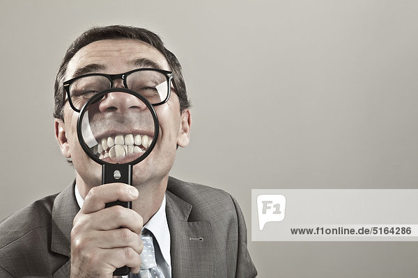 Close up of mature businessman showing teeth through magnifying glass against grey background