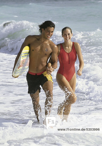 Paar  laufen am Strand durchs flache Wasser  er mit Surfbrett
