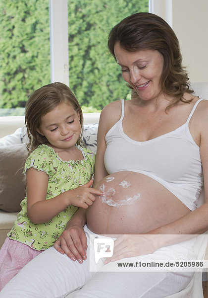 Kleines Mädchen malt mit Creme Gesicht auf den Bauch von schwangerer Mutter