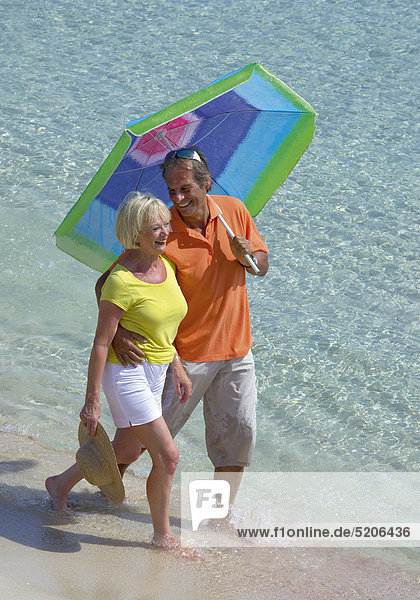 Älteres Paar geht mit Sonnenschirm am Strand spazieren