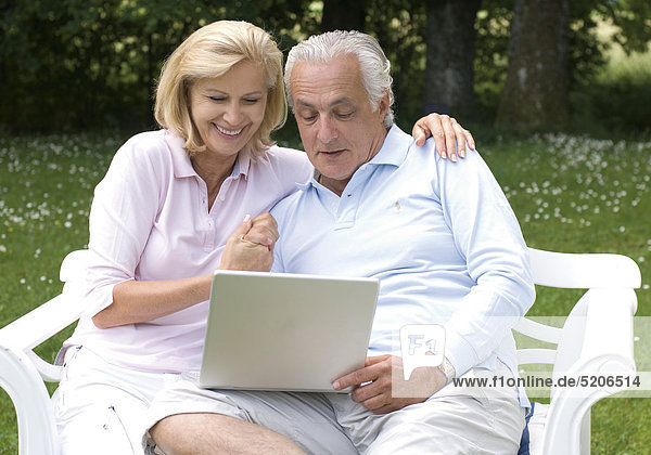 Älteres Paar sitzt im Garten mit Laptop auf Bank