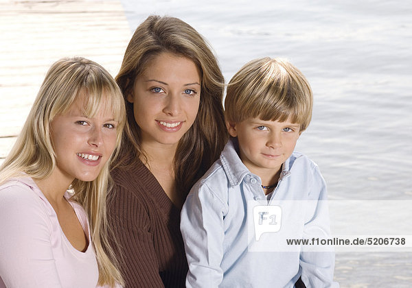 Zwei Teenager-Mädchen und ein kleiner Junge sitzen auf Steg am Wasser