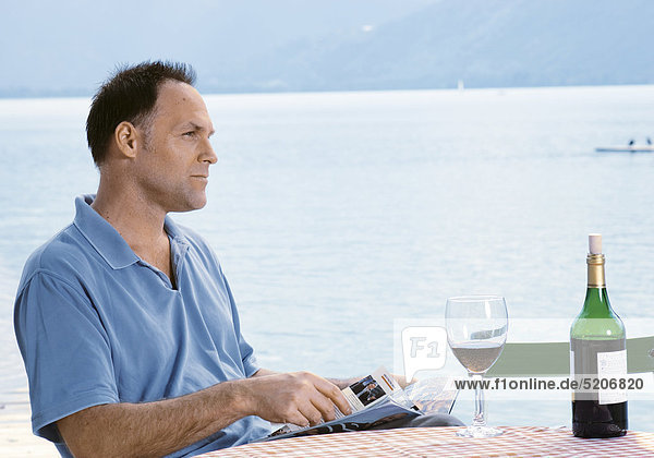 Mann sitzt an See an einem Tisch  liest Zeitschrift  Rotwein