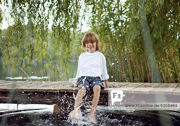 Kleiner Junge sitzt auf Steg und planscht im Wasser