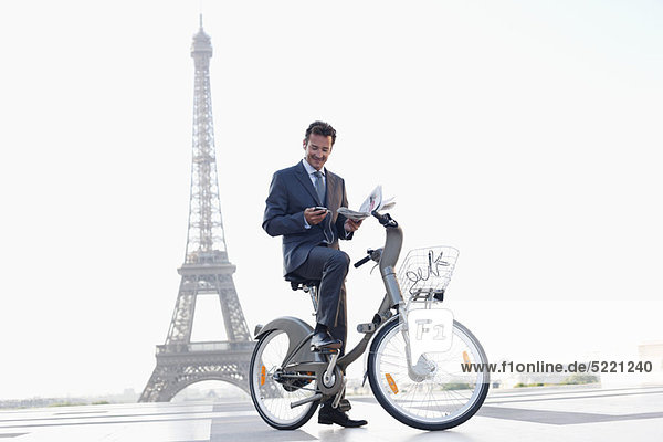 Geschäftsmann mit einer Zeitung und einem Handy auf einem Fahrrad mit dem Eiffelturm im Hintergrund  Paris  Ile-de-France  Frankreich