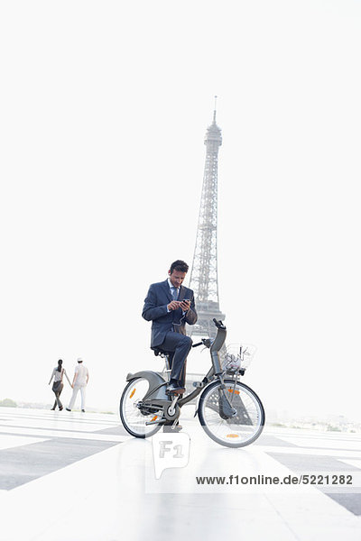 Geschäftsmann  der ein Mobiltelefon auf einem Fahrrad mit dem Eiffelturm im Hintergrund bedient  Paris  Ile-de-France  Frankreich
