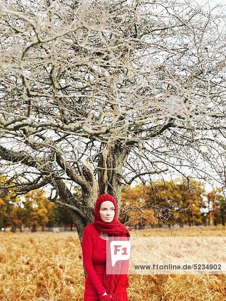 A woman wearing a bright red dress and scarf standing under a leafless tree in nature
