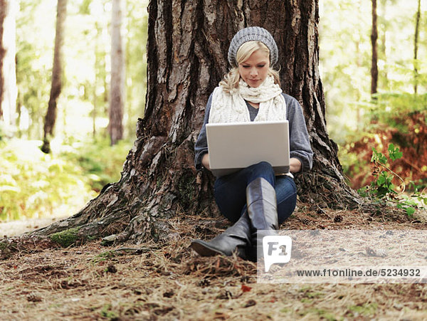A woman in nature leaning against a tree and using a laptop