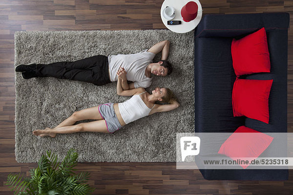 Ein Paar auf einem Wohnzimmerteppich liegend  Händchen haltend und sich gegenseitig anschauend  Blick nach oben