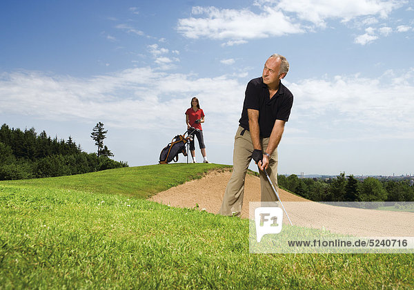 Älterer Mann auf Golfplatz an Sandbunker  junge Fau mit Caddy