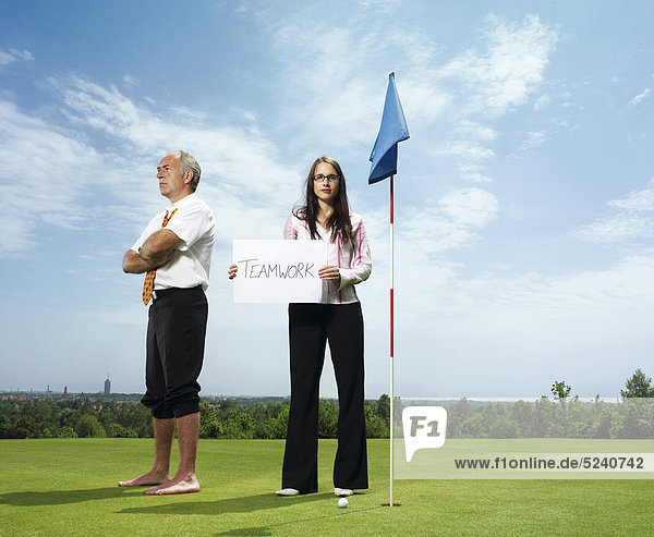Älterer Mann  barfuß und Frau mit Schild Teamwork auf Golfplatz