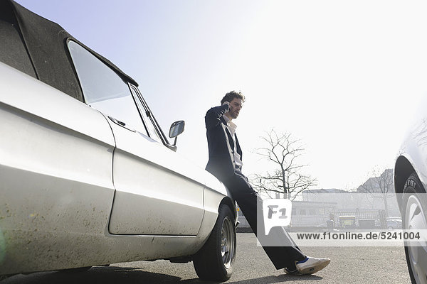 Mittlerer Erwachsener Mann  der sich auf Oldtimer stützt und mit dem Handy spricht