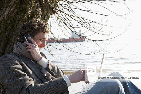 Mann mit Laptop und Handy am Elbufer mit Containerschiff im Hintergrund