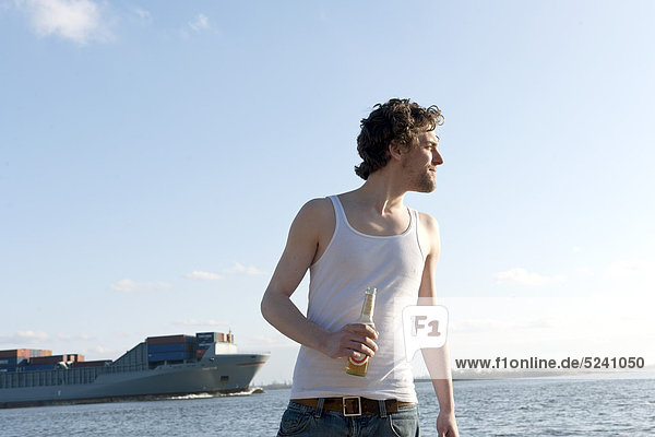Mann mit Bierflasche am Elbufer und Containerschiff im Hintergrund