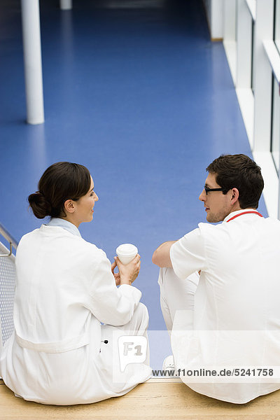 Germany  Bavaria  Diessen am Ammersee  Two young doctors sitting and discussing on staircase with disposable cup  smiling