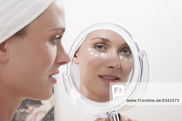 Junge Frau in Handtuch eingewickelt  mit Gesichtscreme und Blick in den Spiegel