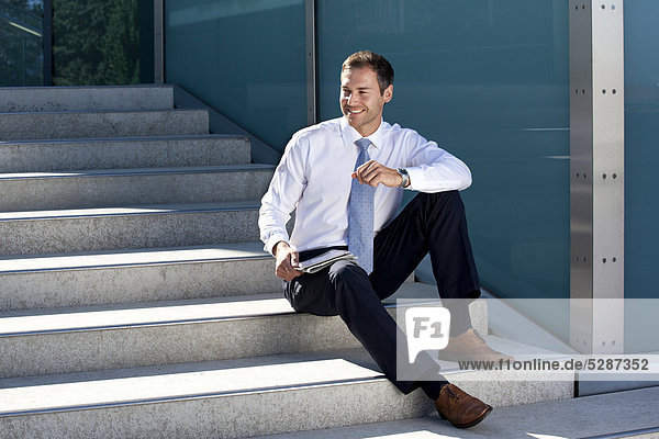 Lächelnder Geschäftsmann sitzt mit Tablet PC und Zeitung auf einer Treppe