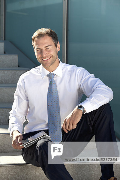 Lächelnder Geschäftsmann sitzt mit Tablet PC und Zeitung auf einer Treppe