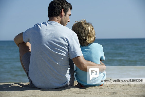Vater und Sohn sitzen zusammen am Strand  Rückansicht
