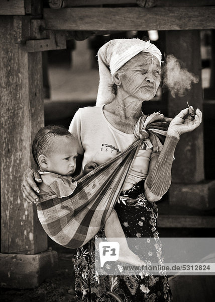 Alte Frau  Großmutter  raucht Zigarre  mit Kind im Tragetuch  in Dorf am Mekong  Laos  Südostasien  Asien