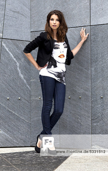 Junge Frau in Top mit Porträtmotiv und Jeans posiert an grauer Wand