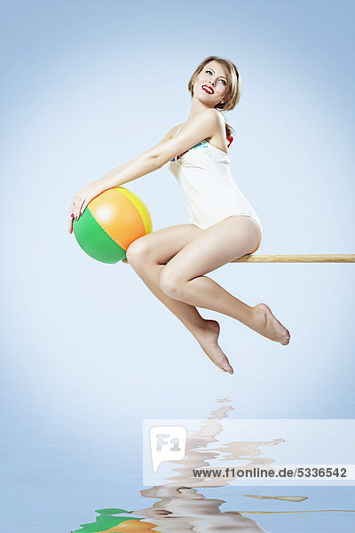 Junge Frau in einem hellen Badeanzug sitzt auf einem Sprungbrett und hält einen Wasserball  Pin-up