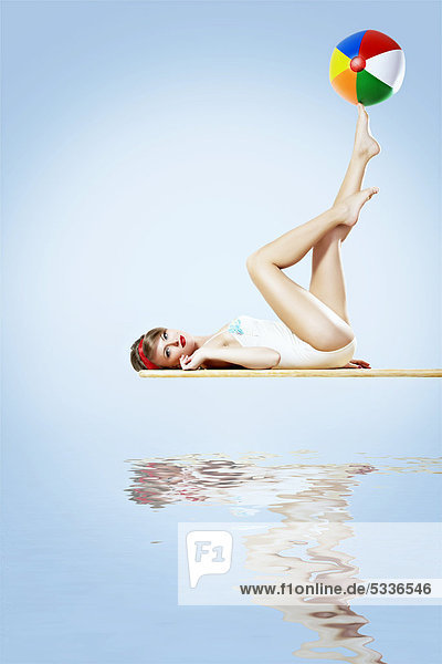 Junge dunkelblonde Frau in einem hellen Badeanzug liegt auf einem Sprungbrett  einen Wasserball auf den Zehenspitzen balancierend  Pin-up