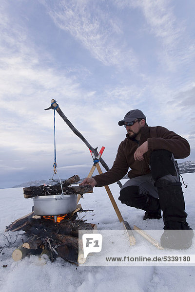 Mann kocht mit Lagerfeuer und hängendem Anheizherd oder Dutch Oven  Eis auf zugefrorenem Lake Laberge  Yukon Territory  Kanada