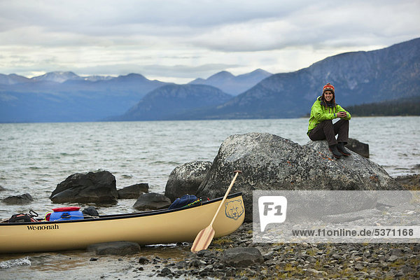 Junge Frau sitzt auf einem Felsen  Kanu und Holzpaddel am Ufer  Kusawa Lake  die Berge dahinter  Yukon Territory  Kanada