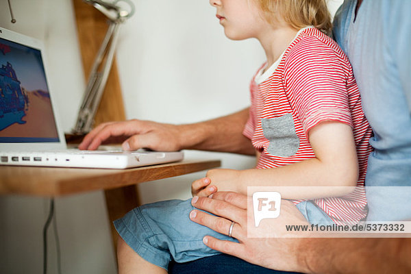 Boy sitting on father's lap at computer desk