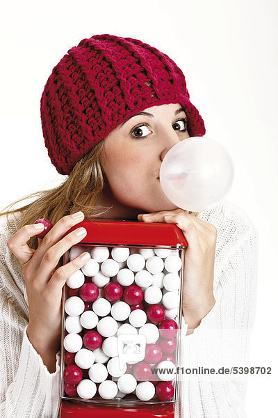 Young woman wearing a red woolen cap blowing a bubble with bubble gum behind a gumball machine