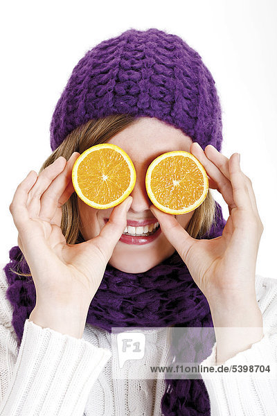 Young woman wearing a purple woolen cap and scarf holding orange halves in front of her eyes