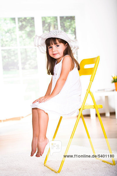 Young girl wearing a white dress and hat  sitting on a chair